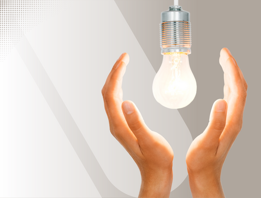 Ein Paar Hände mit nach innen gerichteten Handflächen befindet sich unter einer leuchtenden Glühbirne, die aus einer Fassung hängt, vor einem hellgrauen und weißen Hintergrund.