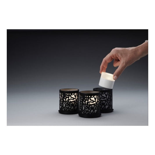 Eine Hand hebt eine kleine weiße LED-Kerze aus einer schwarzen Halterung, während zwei identische Duni GmbH Multicolour LED Set, Mini - 8 Stück in ähnlichen Halterungen auf einer grauen Fläche vor dunklem Hintergrund ruhen.