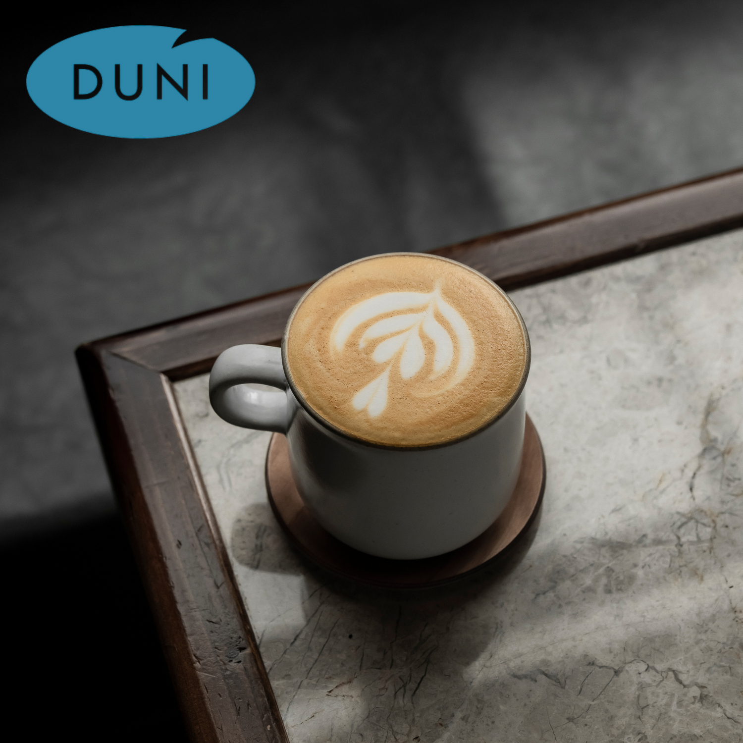 Eine Tasse Kaffee mit Latte-Art, serviert auf einem Holzuntersetzer auf einem Marmor-Tisch, begleitet vom Duni-Logo im oberen linken Bereich