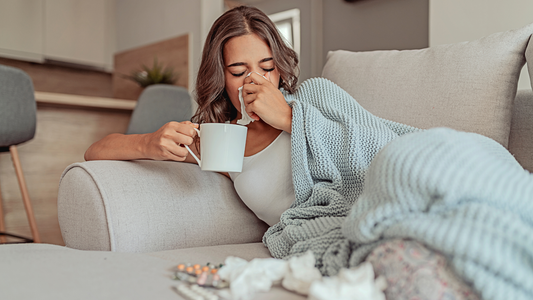 Frau, die auf dem Sofa liegt und eine Tasse in der einen Hand hält, während sie sich mit der anderen die Nase putzt