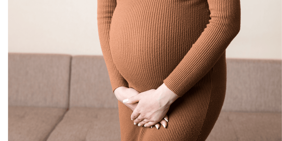 Schwangere Frau in engem, braunem Kleid hält ihren Bauch mit beiden Händen, Nahaufnahme im Innenraum auf Sofa Hintergrund