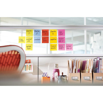 A series of 3M Post-it® Notes, Yellow, 76x76 mm (90 sheets/block, 6 blocks/pack) is arranged on a glass divider; below, on a desk are folders, books, a water bottle, and other utensils.