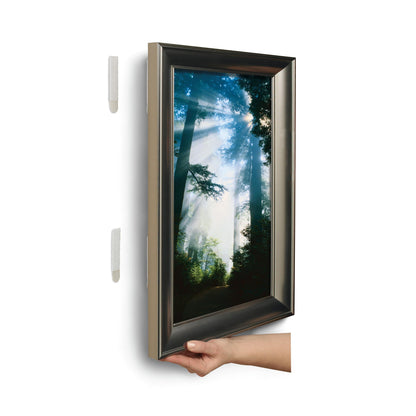 Eine Hand hebt ein gerahmtes Bild von einer weißen Wand und enthüllt zwei 3M Deutschland GmbH Große Command™ Bilder-Montage Strips L, 6 Sets im Vorteilspack, hinter dem Bild.