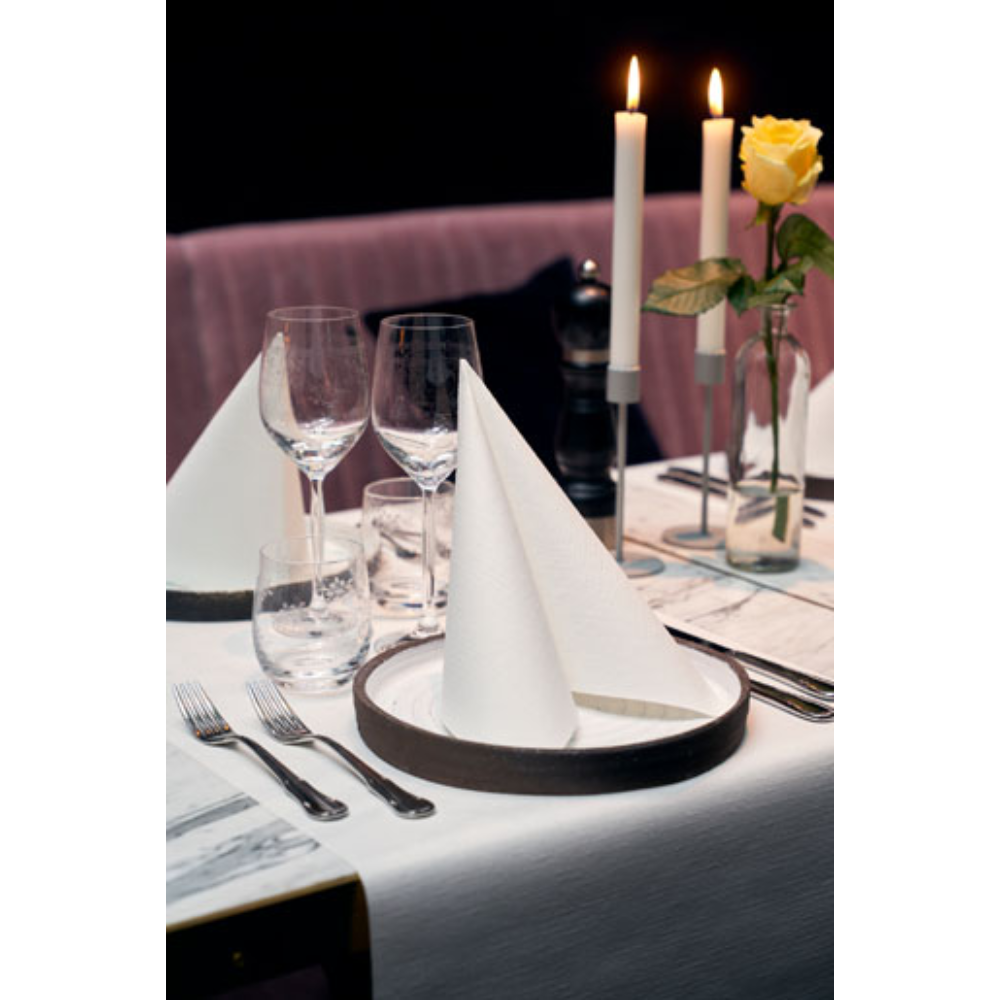 A formal table arrangement includes neatly folded Dunilin® napkins 40 x 40 cm from Duni GmbH on each plate, complemented by wine glasses, a water glass, cutlery, burning candles, and a single yellow rose in a vase. A light pink fabric serves as the background.