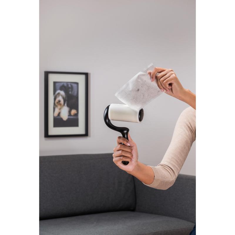 A person peels off a used sheet from a Scotch-Brite™ Everyday Clean Lint Roller for flat surfaces (56 sheets, 1/package) by 3M Deutschland GmbH while standing in front of a gray sofa and a photo of a dog - ideal for cleaning flat surfaces.