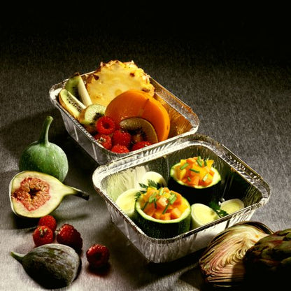Two Starpak 25 aluminum trays with PP-coated lids are filled with fresh fruits like kiwi, pineapple, figs, berries, and vegetables like zucchini and melon on a gray surface - ideal for catering or take-away.