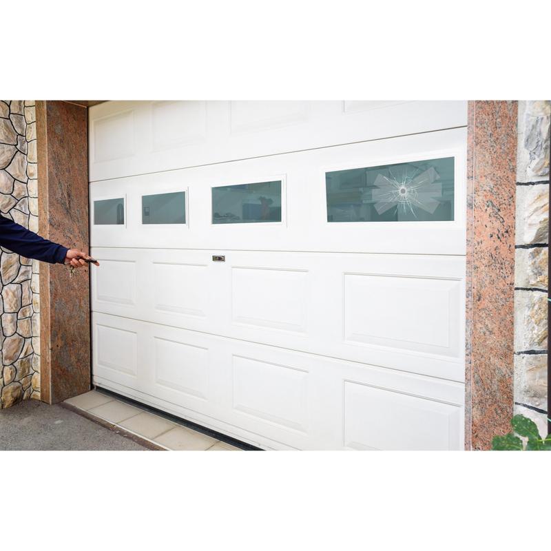 A person points to a white garage door with four rectangular windows, one of which is broken in a spider web pattern - ideal for repair with 3M Deutschland GmbH Scotch® Extremium™ INVISIBLE High-Performance Tape (20 m x 48 mm). Stone and tiles border the garage.