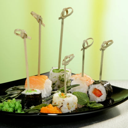 On a black plate lie various sushi rolls, each skewered with a PAPSTAR 250 Fingerfood - Picker "pure" of PAPSTAR GmbH, arranged with green garnishes against a blurred yellow-green background.