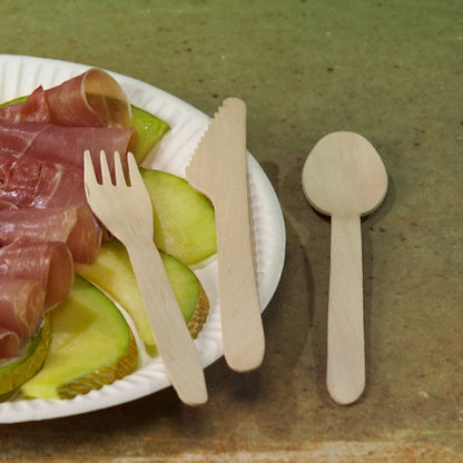 A white paper plate with green apple slices and prosciutto is on a table. Next to it are PAPSTAR wooden spoons "pure" 15.7 cm - eco-friendly, sturdy wooden spoons (100 pieces), from PAPSTAR GmbH, neatly arranged on the brown surface.