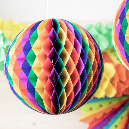 A close-up of the PAPSTAR flame-retardant honeycomb ball from PAPSTAR GmbH, a spherical decoration in rainbow colors, with other colorful decor gently blurred in the background.