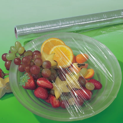 A bowl with various fruits is covered with perforated Starpak cling film, while an unused roll of Starpak lies behind it on a green surface - perfect for keeping food fresh.