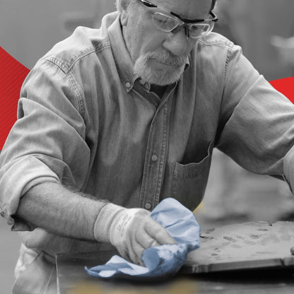 An older man in a denim shirt uses a blue WypAll® L20 paper towel for industrial cleaning tasks from Kimberly-Clark GmbH. He wears safety glasses and gloves while wiping a metal surface; the background has red and yellow accents.