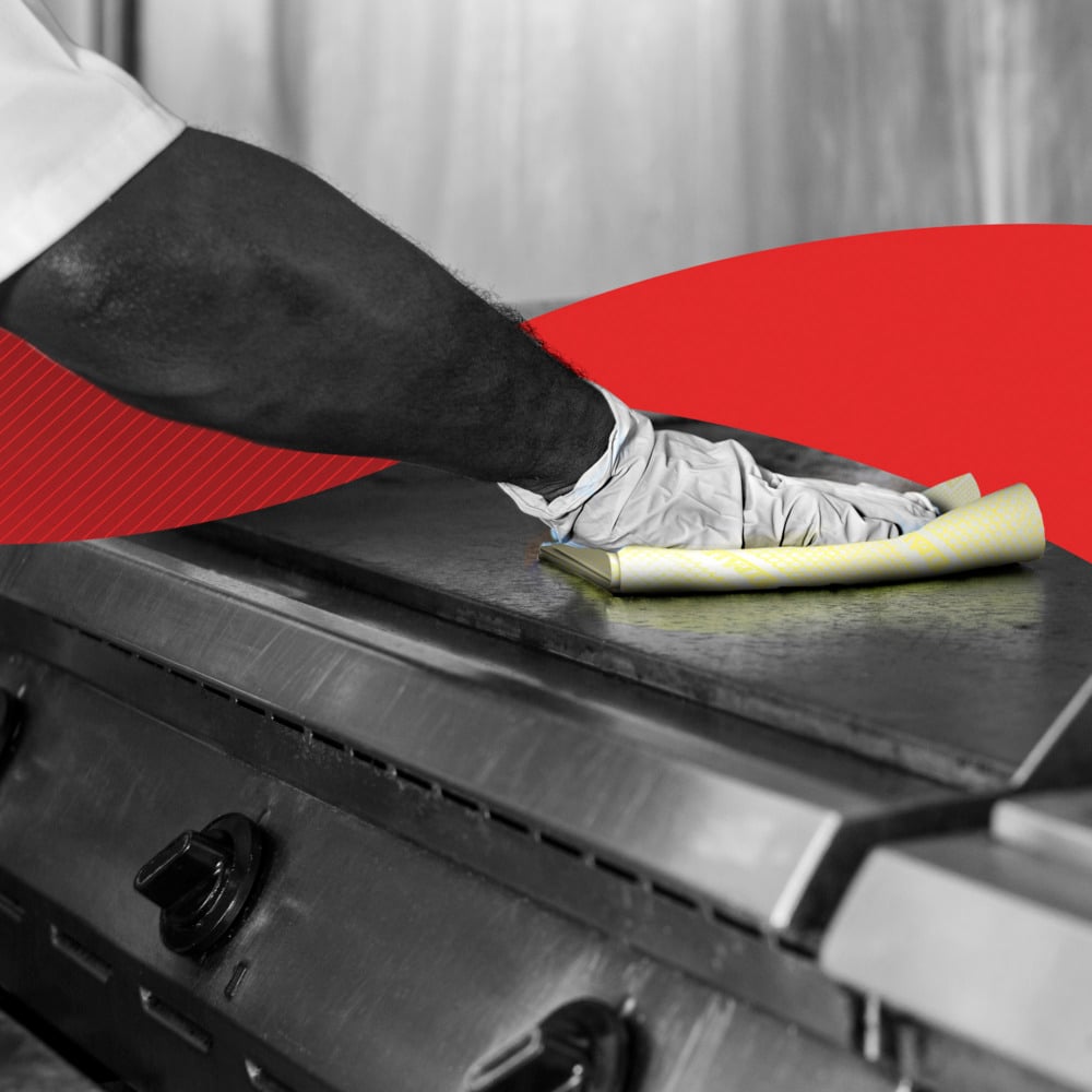 Eine Person wischt mit Handschuhen einen gewerblichen Küchenherd mit einem gelben WypAll® X80 Critical Clean - Interfold (41 x 33 cm) der Kimberly-Clark GmbH ab; im Hintergrund erscheint ein rotes Grafikelement.