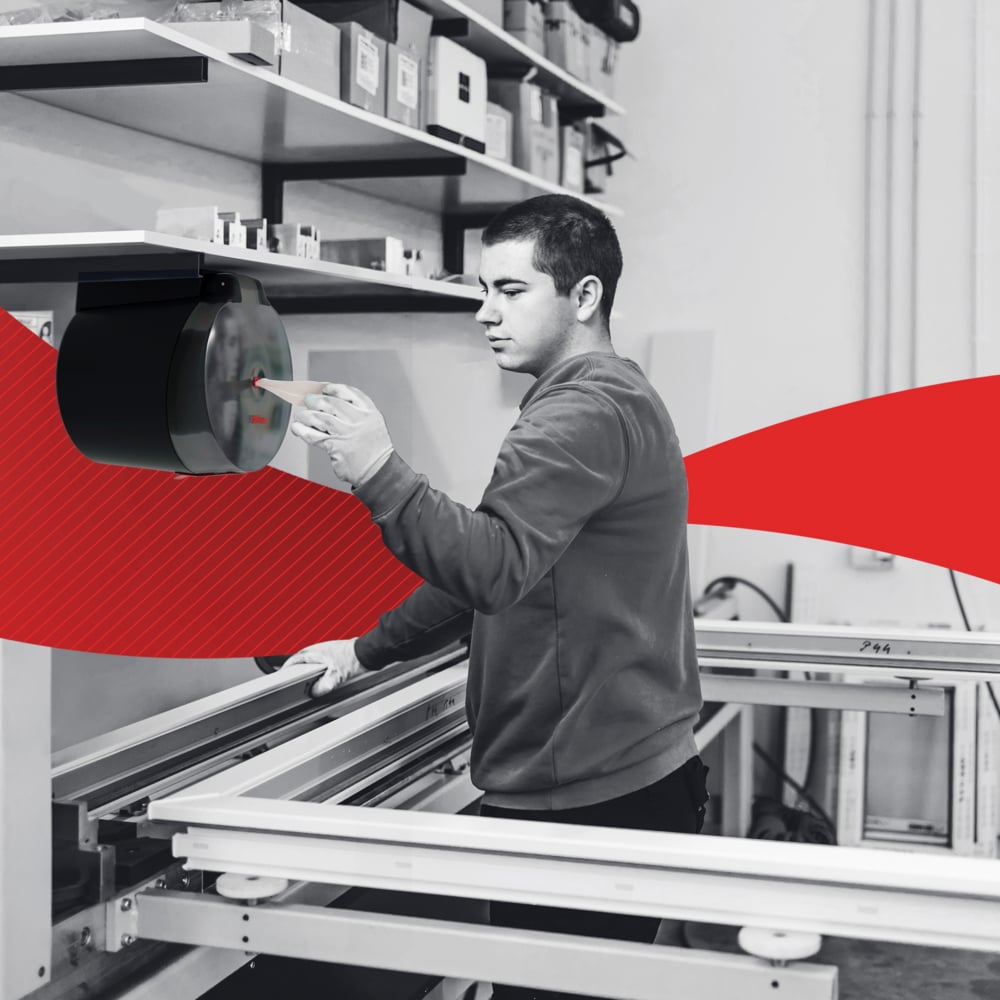 In a workshop, a person extracts a WypAll® L20 paper towel from Kimberly-Clark GmbH via central dispensing from the wall dispenser and cleans industrial work surfaces; a red graphic element runs through the center of the image.