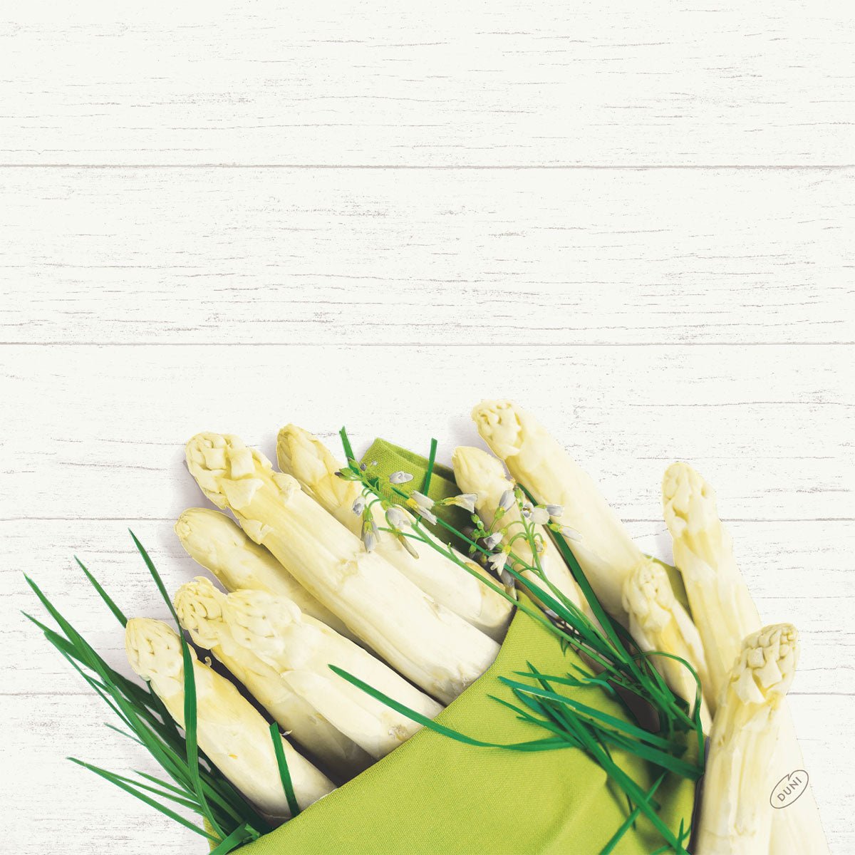 A bundle of white asparagus and green herbs rests on a 40x40cm Bio-Dunisoft napkin from Duni GmbH against a white wooden background.
