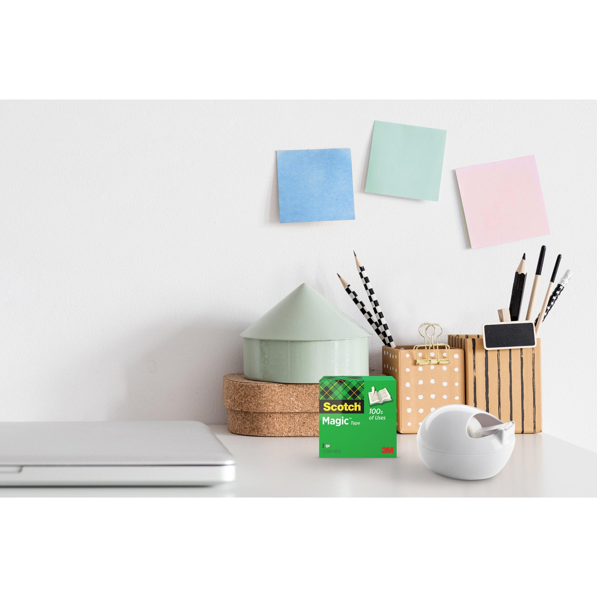 Ein Schreibtisch mit einem geschlossenen Laptop, ein 3M Deutschland GmbH Scotch™ Karim Rashid Tischabroller und eine Schachtel Klebeband, Behälter für Stifte und Bleistifte, ein kleines Ornament in Form eines Hauses und drei bunte Klebezettel an der weißen Wand.