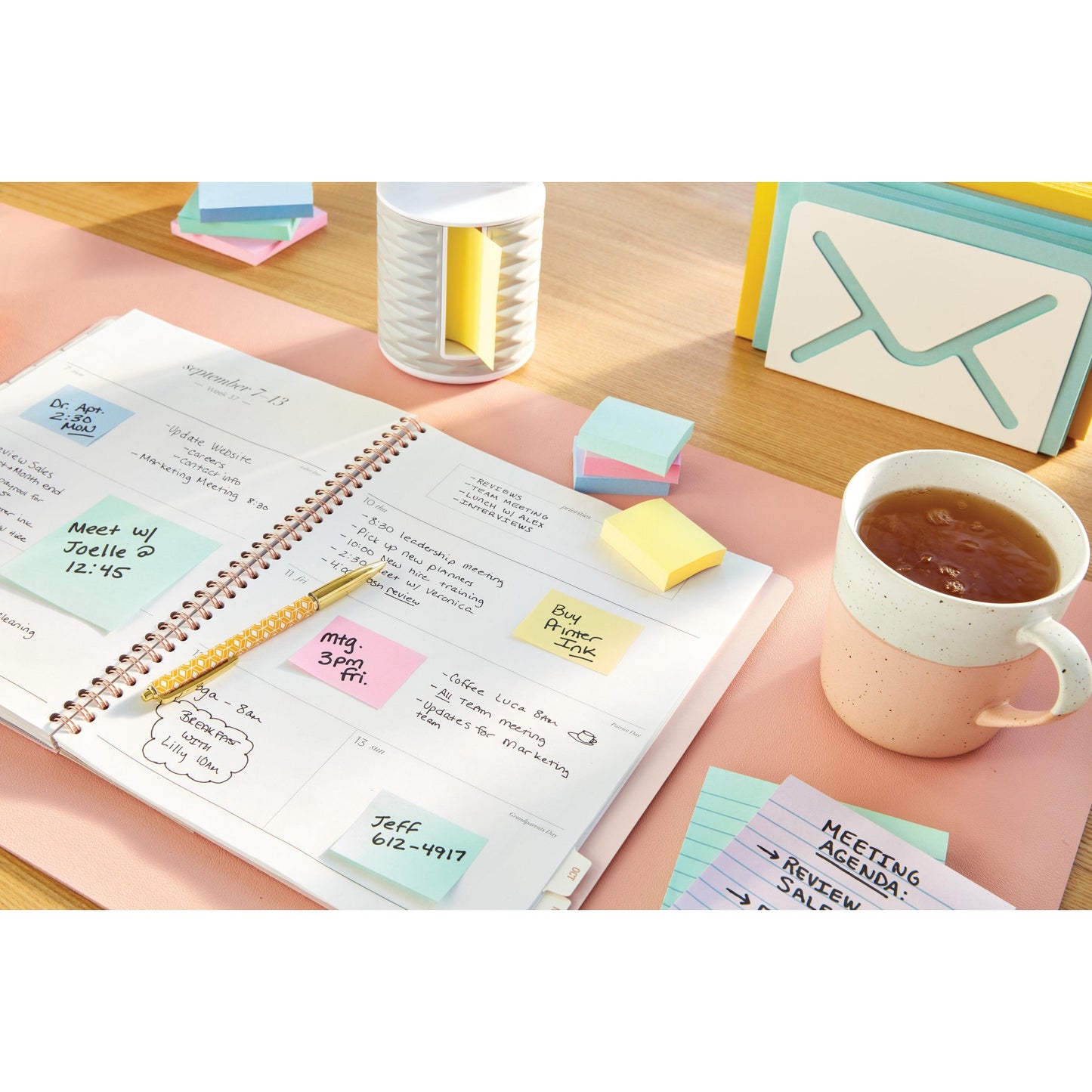 Ein offener Planer mit handschriftlichen Notizen liegt auf einem Schreibtisch neben einer Tasse Tee, Karteikarten, einem Stift und Post-it® Notes in Gelb (38 mm x 51 mm, 3M Deutschland GmbH) sowie einem Behälter.