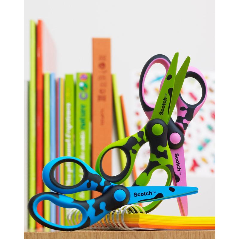Two Scotch™ DECO children's scissors (green, blue, or pink, 13 cm) from 3M Deutschland GmbH with colorful patterns and rounded tips lie on a stack of paper. In the background, there are colorful books.