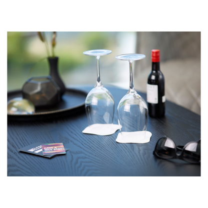 On a wooden table, there are two upside-down wine glasses with white, multi-layered coasters from Duni GmbH underneath. A small wine bottle, a pair of black sunglasses, and a tray full of decorative items add charm. Next to it, a 'Live & Life Safe' card waits to provide inspiration.