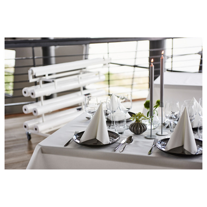 Auf einem hübsch gedeckten Esstisch steht eine frische, weiße Duni Dunicel®-Tischdeckenrolle der Duni GmbH, die eine stoffähnliche Haptik bietet. Dreieckig gefaltete Servietten auf dunklen Tellern begleiten Weingläser. Hohe Kerzen und kleine Blumenarrangements dienen als Tischdekoration, im Hintergrund sind gerollte Leinwände zu sehen.