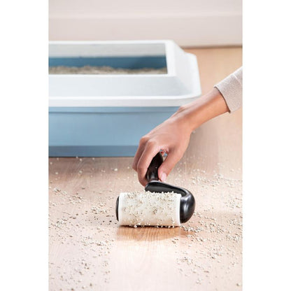 A hand uses the Scotch-Brite™ Everyday Clean Lint Roller for flat surfaces (56 sheets, 1/package) from 3M Deutschland GmbH to pick up scattered cat litter from a wooden floor, with a cat litter box with a blue lid visible in the background.