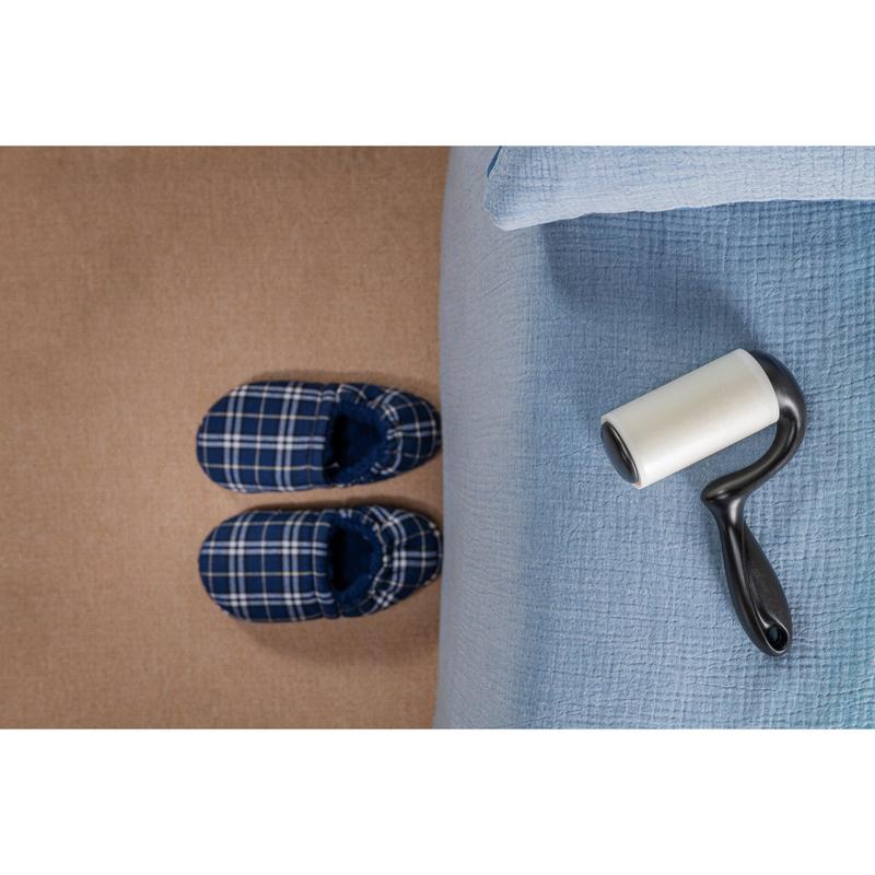 A pair of blue checkered slippers stands on a beige carpet next to a bed with a light blue bedspread, while the 3M Deutschland GmbH Scotch-Brite™ Everyday Clean Lint Roller for flat surfaces (56 sheets, 1/package) lies on the bed ready for use.