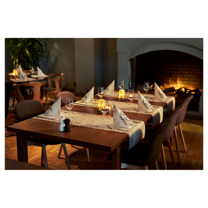 Ein gemütlicher Essbereich verfügt über zwei Holztische mit Weingläsern und Duni Bio Dunisoft® Servietten neben flackernden Kerzen. Dunkle Stühle umgeben die Tische, aufgewertet durch einen brennenden Kamin. Plüschige Sitzgelegenheiten und eine Pflanze tragen zum naturinspirierten Design bei.