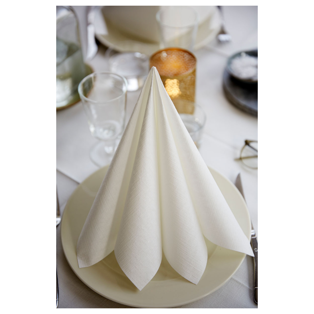A neatly folded Duni Dunilin® napkin 48 x 48 cm White by Duni GmbH stands upright on a white plate amidst empty glasses, a round candle holder, a salt bowl, cutlery, and a white tablecloth. The elegant ambiance highlights the fabric-like texture of the high-quality disposable napkin.