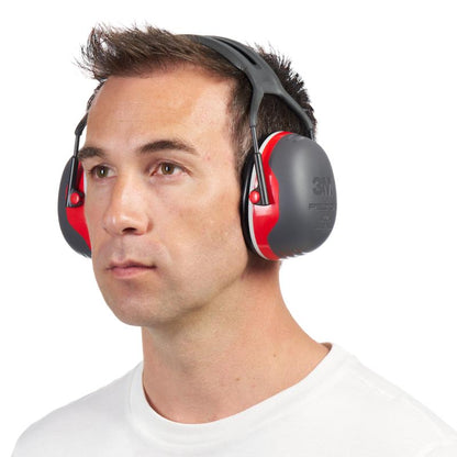 A man in a plain white shirt wears gray and red 3M™ PELTOR™ Comfort Earmuffs from 3M Deutschland GmbH and gazes forward with a neutral facial expression against a white background.