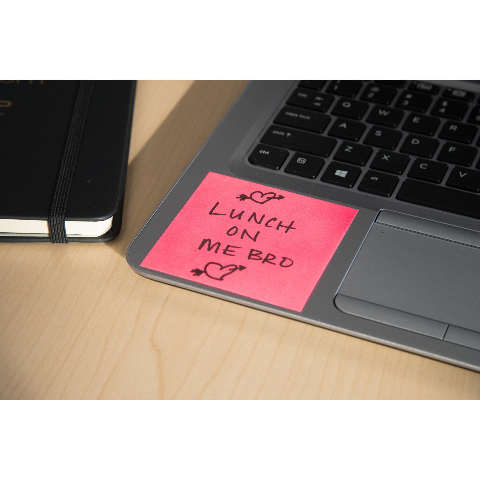 Ein Post-it® Notes Würfel (76x76mm, 450 Blatt, 100% PEFC) von 3M Deutschland GmbH in Pink sitzt auf einer Laptop-Tastatur mit "LUNCH ON ME BRO" und Herz-Kritzeleien. Ein geschlossenes schwarzes Notizbuch liegt daneben auf einem hellen Holztisch.