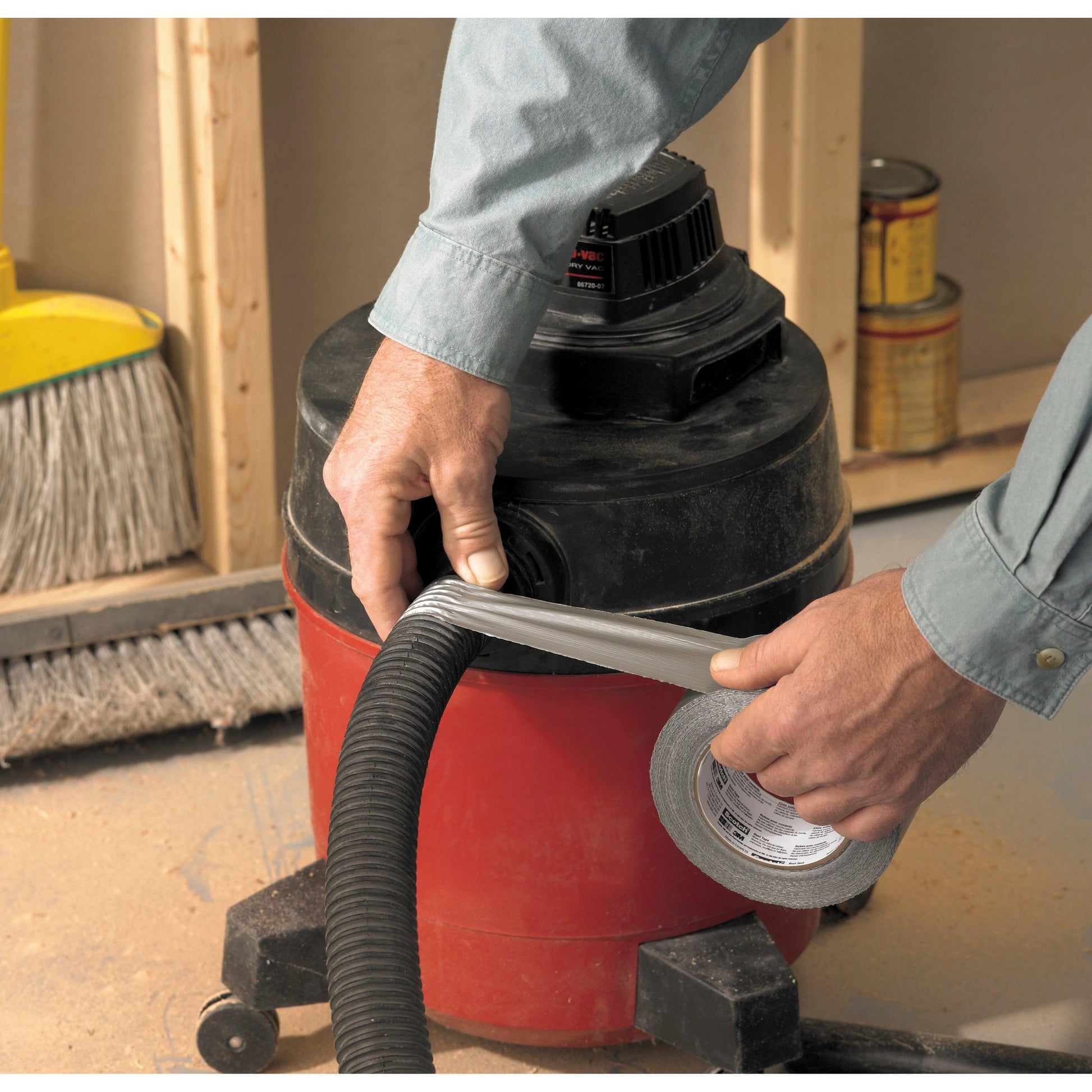 Jemand wickelt Scotch® Extremium™ Universal Klebeband (3 m x 19 mm) von 3M Deutschland GmbH um den Schlauch eines rot-schwarzen Werkstattsaugers. Ein Besen und Farbdosen liegen auf dem staubigen Boden einer Baustelle.