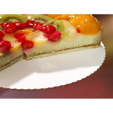 On a round, wavy PAPSTAR 10 cake board, cardboard "pure" by PAPSTAR GmbH, lies a sliced cake with fruit topping, decorated with cherries, kiwi, and shiny peach slices; a part of the cake has been removed.