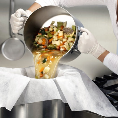 Dressed in white gloves, someone pours vegetables and broth through parchment-lined straining and filter cloths (100x100 cm, 10 pieces) by Starpak into a large metal container, thus demonstrating practical use in the kitchen.