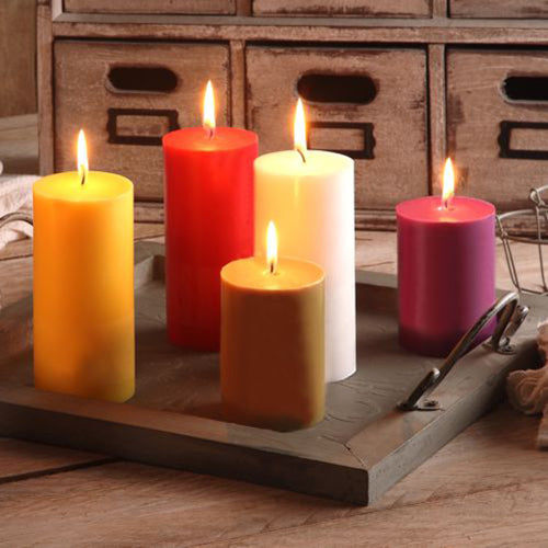 Five burning PAPSTAR pillar candles Ø 70 mm, 100 mm made of 100% stearin, dyed candles in yellow, red, white, orange, and purple from PAPSTAR GmbH are arranged on a wooden tray with handles in front of a rustic drawer unit.