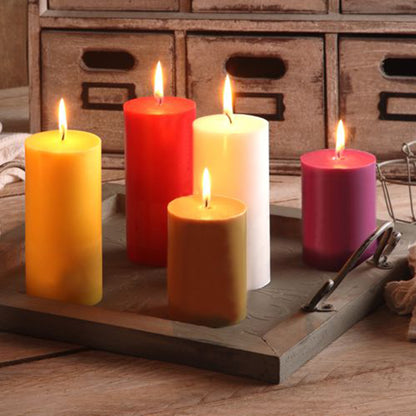 Five burning PAPSTAR pillar candles Ø 70 mm, 100 mm made of 100% stearin, dyed candles in yellow, red, white, orange, and purple from PAPSTAR GmbH are arranged on a wooden tray with handles in front of a rustic drawer unit.