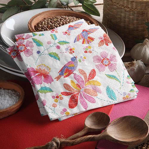 A stack of PAPSTAR 20 napkins, 25 x 25 cm, 3-ply, 1/4-fold from PAPSTAR GmbH with colorful flower and bird motifs lies on a table between wooden spoons, grains, and a basket – perfect as a vibrant table decoration. Plates in the background.