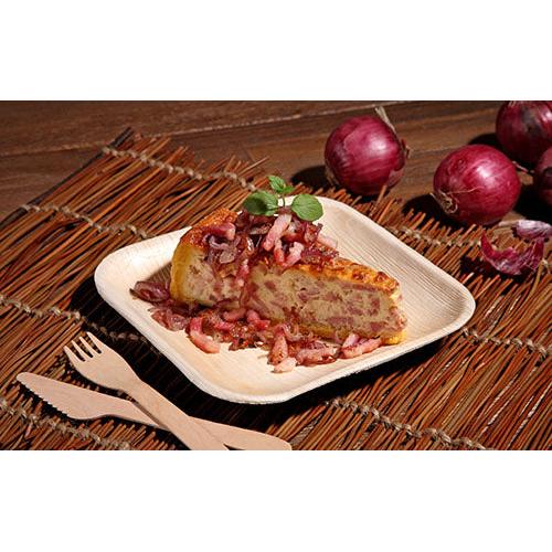 A slice of Spanish tortilla with onions and herbs is served on a square plate PAPSTAR 25, palm leaf "pure" from PAPSTAR GmbH, resting on a woven mat with wooden cutlery and red onions in the background.