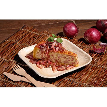 A slice of Spanish tortilla with onions and herbs is served on a square plate PAPSTAR 25, palm leaf "pure" from PAPSTAR GmbH, resting on a woven mat with wooden cutlery and red onions in the background.