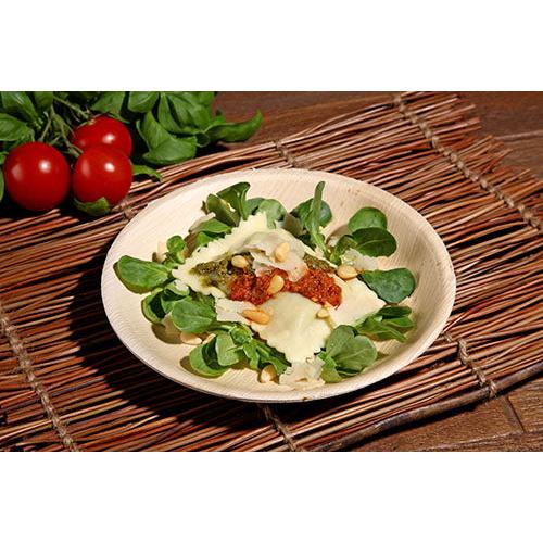 A plate of ravioli with tomato sauce, pine nuts, and cheese on leafy greens is served on a PAPSTAR 25 plate palm leaf "pure" round from PAPSTAR GmbH, resting on a bamboo placemat with cherry tomatoes and leafy greens in the background.