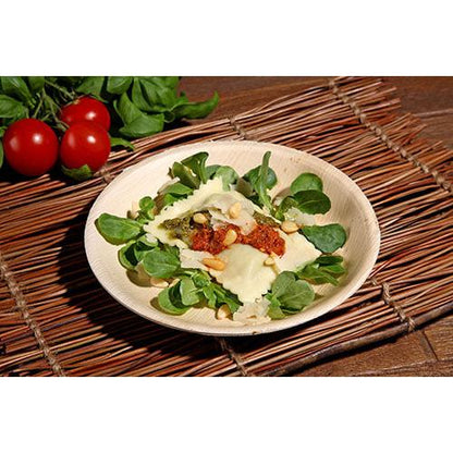 A plate of ravioli with tomato sauce, pine nuts, and cheese on leafy greens is served on a PAPSTAR 25 plate palm leaf "pure" round from PAPSTAR GmbH, resting on a bamboo placemat with cherry tomatoes and leafy greens in the background.