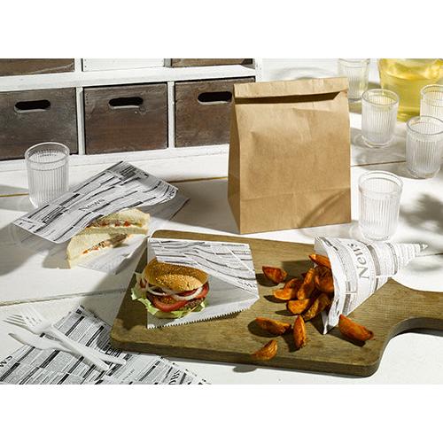 A sandwich, a burger, and seasoned wedges are served with clear cups, plastic cutlery, napkins, and PAPSTAR 5 wrapping papers "Newsprint" (3cm x 2cm) from PAPSTAR GmbH on a wooden board next to a brown paper bag.