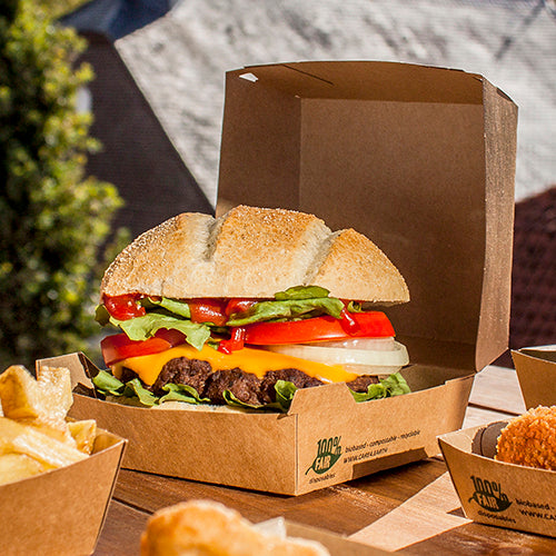 Auf einem Outdoor-Tisch liegt ein Hamburger mit Pommes in einem umweltfreundlichen PAPSTAR 25 Burgerbox aus brauner Pappe (9 x 18,5 x 18,5 cm, „100% Fair“, extra groß) von PAPSTAR GmbH.