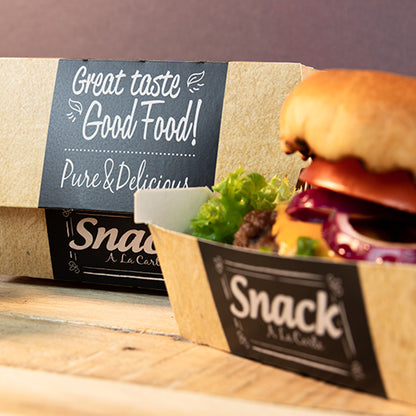 Ein Papptablett mit der Aufschrift "Snack A La Carte" enthält einen Hamburger mit Salat, Tomate und Zwiebel. Im Hintergrund steht eine PAPSTAR 50 Burgerboxen, Pappe "pure" "Good Food" von PAPSTAR GmbH: Guter Geschmack Good Food! Pur & Lecker.