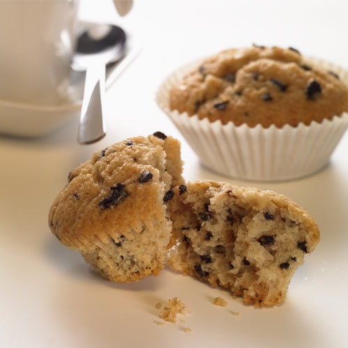 Ein Schokoladenmuffin liegt in zwei Hälften auf einer weißen Fläche, daneben ein ganzer Muffin in einem runden 1000er Backförmchen der PAPSTAR GmbH, im Hintergrund eine Tasse mit Löffel. Neben dem halbierten Muffin sind Krümel verstreut.