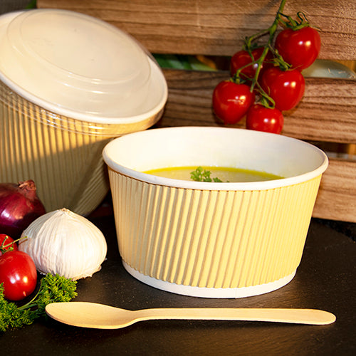 A PAPSTAR round soup cup, brown/white, Ripple Wall, 720 ml (25 pieces) from PAPSTAR GmbH contains yellow soup with herbs, alongside a wooden spoon, garlic, red onion, vine tomatoes, and green parsley.