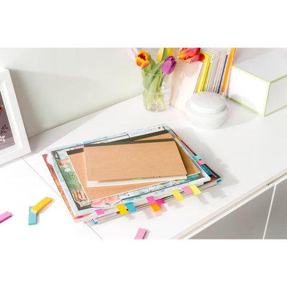 On a white desk lies the Post-it® Page Marker (15 mm x 50 mm, 50 sheets/block, 100% PEFC) from 3M Deutschland GmbH between stacked books and notebooks with colorful markers, scattered notes, a glass vase with flowers, and stationery.