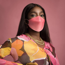 Eine Person trägt eine partikelfiltrierende Altruan Designer-Gesichtsmaske (FFP2-Standard) in einem stilvollen Rosaton und ein farbenfrohes Oberteil mit abstraktem Muster vor einem rosa Hintergrund. Sie hat langes, dunkles Haar und trägt mehrere goldene Halsketten.