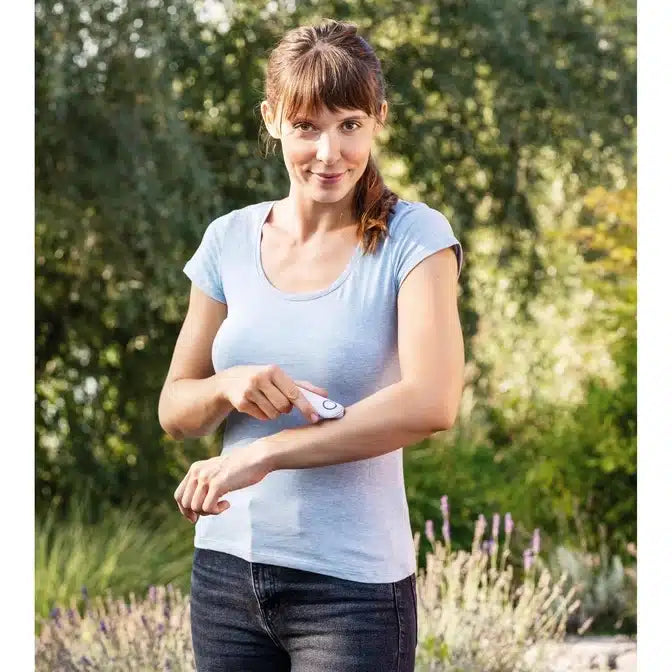 A woman with brown hair, wearing a white T-shirt and black jeans, applies an insect bite healer BR 10 from Beurer GmbH to her right upper arm outdoors. In the background, green leaves and lavender plants can be seen.