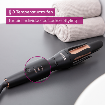A black Beurer HT 75 automatic curling iron with rose gold accents stands on a white surface next to neatly rolled gray towels. The German text on the image reads: "3 temperature settings for customized curling styling." The brand name "Beurer GmbH" is visible on the curling iron.