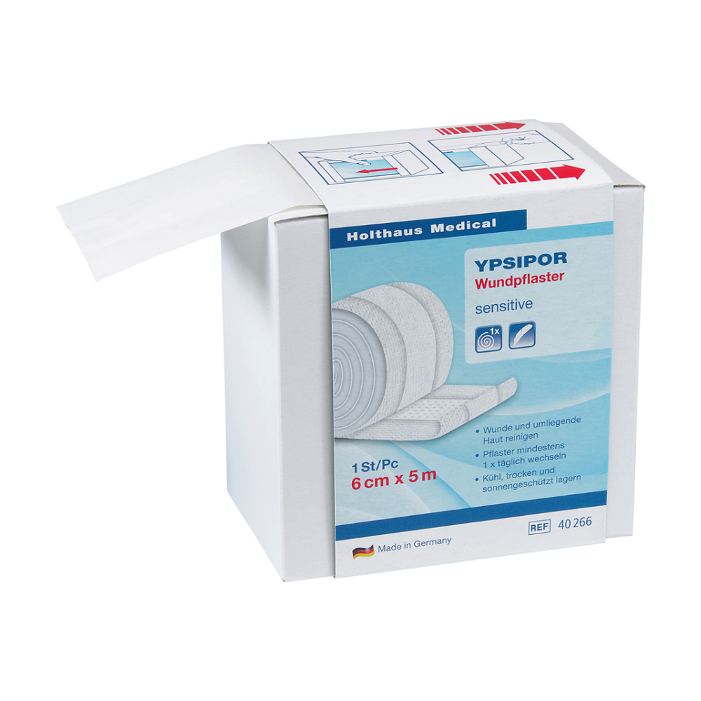 Tá bosca de phlaistéir créachta Holthaus Ypsipor Vlies ó Holthaus Medical GmbH & Co. KG le feiceáil. Tá sé scríofa ar an mbosca go bhfuil sé íogair, go bhfuil sé 6 cm x 5 m, agus go bhfuil sé déanta sa Ghearmáin. Tá cuid den rolla créachta craiceann-chairdiúil le feiceáil agus ag gobadh amach as an mbosca.